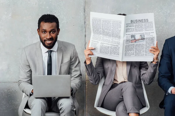 Lächelnder afrikanisch-amerikanischer Geschäftsmann mit Laptop, während Geschäftsfrau Zeitung in Wartehalle liest — Stockfoto