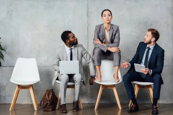 Hommes d'affaires multiethniques utilisant des appareils numériques tandis que la femme d'affaires grave assis sur une chaise avec les bras croisés dans la salle d'attente — Photo de stock