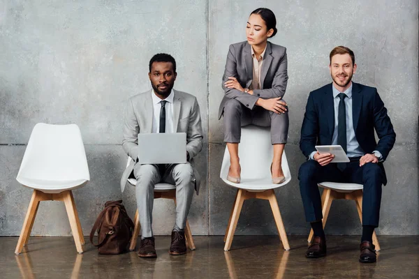 Multiethnische Geschäftsleute benutzen digitale Geräte, während Geschäftsfrau mit verschränkten Armen im Wartezimmer auf einem Stuhl sitzt — Stockfoto