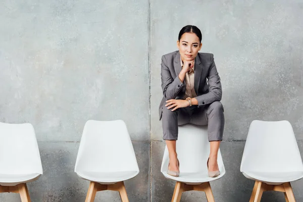 Beautiful asian businesswoman in formal wear sitting on chair and looking at camera in waiting hall — Stock Photo