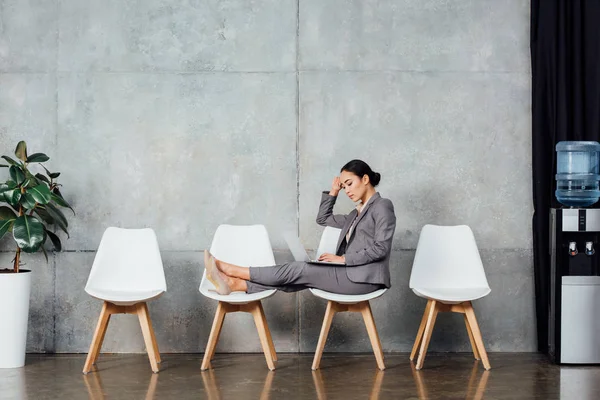 Pensive asian businesswoman in formal wear sitting on chairs and using laptop in waiting hall — Stock Photo