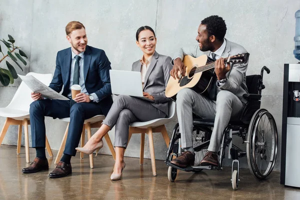 Empresarios multiculturales sentados y escuchando mientras el hombre afroamericano discapacitado toca la guitarra en la sala de espera - foto de stock