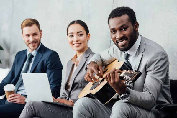 Empresarios multiculturales sentados y escuchando mientras el hombre afroamericano toca la guitarra en la sala de espera - foto de stock