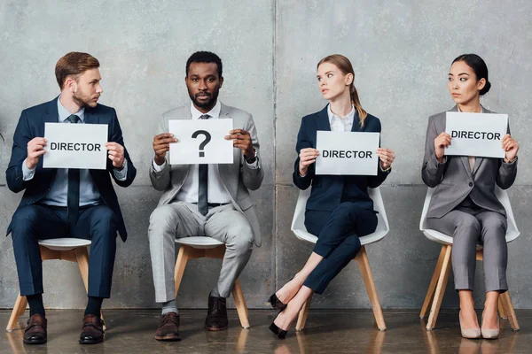 Multiethnic businesspeople sitting and holding cards with 'director' words and question mark in waiting hall — Stock Photo