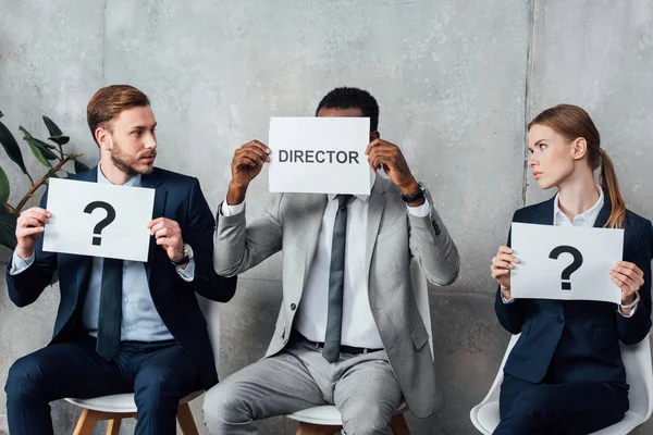 Multiethnic businesspeople holding cards with 'director' word and question marks in waiting hall — Stock Photo