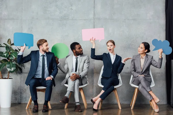 Multiethnic businesspeople holding speech bubbles and thought bubble while sitting in waiting hall — Stock Photo