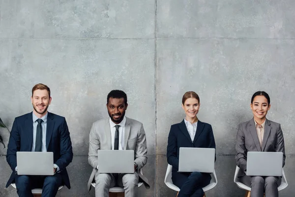 Happy multiethnic businesspeople sitting on chairs and using laptops in waiting hall — Stock Photo