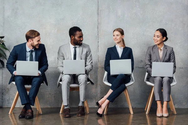 Sonrientes empresarios multiétnicos sentados en sillas y usando computadoras portátiles en la sala de espera - foto de stock