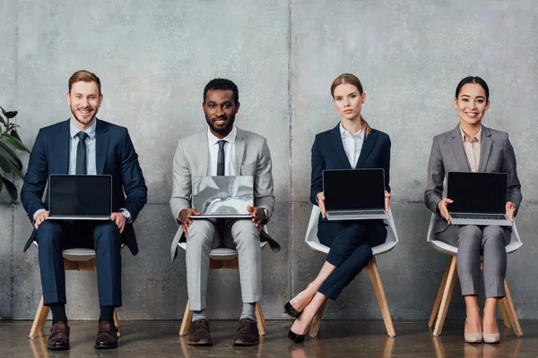 Multiethnic businesspeople sitting on chairs and holding laptops with blank screen in waiting hall — Stock Photo