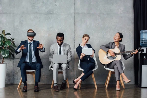 Empresários multiétnicos sentados, usando dispositivos digitais e tocando guitarra acústica na sala de espera — Fotografia de Stock