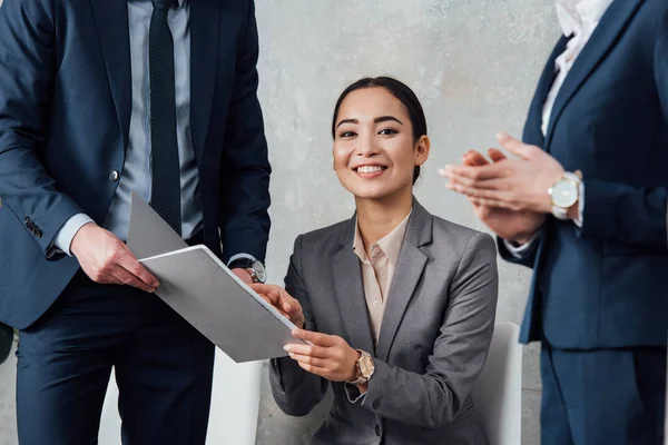 Smiling asian businesswoman holding folder during meeting with colleagues in office — Stock Photo