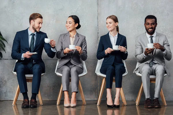 Smiling multiethnic businesspeople sitting on chairs and drinking coffee in waiting hall — Stock Photo