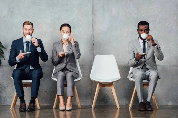 Мультиэтнические предприниматели пьют кофе, сидя на стульях в зале ожидания — стоковое фото
