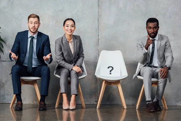 Multiethnic businesspeople sitting near card with question mark on chair in waiting hall and looking at camera — Stock Photo