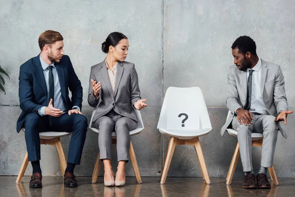 Surpreendidos empresários multiétnicos olhando para o cartão com ponto de interrogação na cadeira no salão de espera — Fotografia de Stock