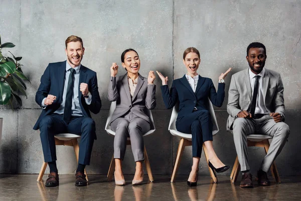 Happy multiethnic businesspeople sitting on chairs and cheering with clenched fists in waiting hall — Stock Photo