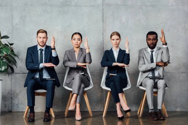Serious multiethnic businesspeople sitting on chairs with raised hands ready to answer in waiting hall — Stock Photo