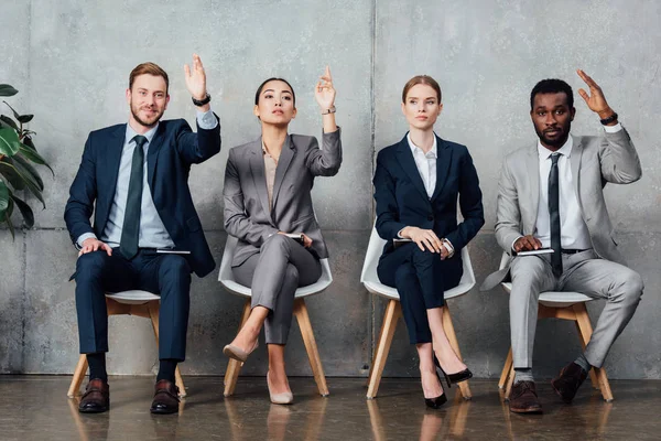 Multiethnic businesspeople sitting on chairs with raised hands ready to answer in waiting hall — Stock Photo