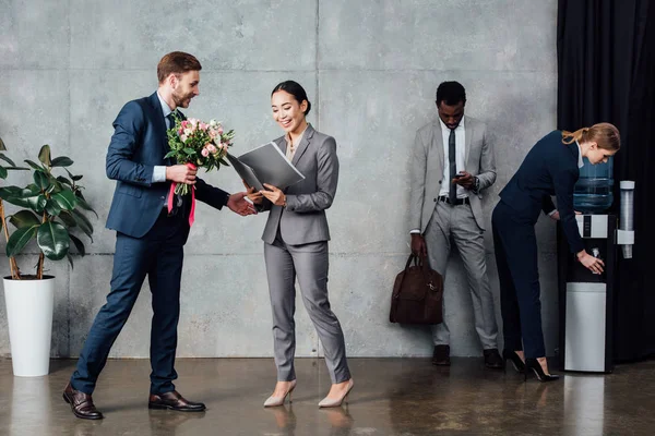 Бизнесмен вручает цветы улыбающейся бизнесвумен с многонациональными коллегами на заднем плане в зале ожидания — стоковое фото