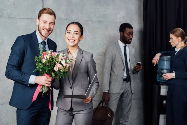 Homme d'affaires souriant présentant des fleurs à une femme d'affaires avec des collègues multiethniques sur fond dans la salle d'attente — Photo de stock