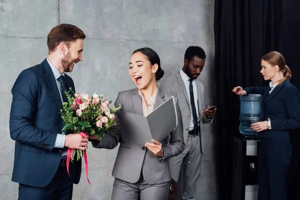 Hombre de negocios presentando flores a alegre mujer de negocios asiática con colegas en fondo en sala de espera - foto de stock