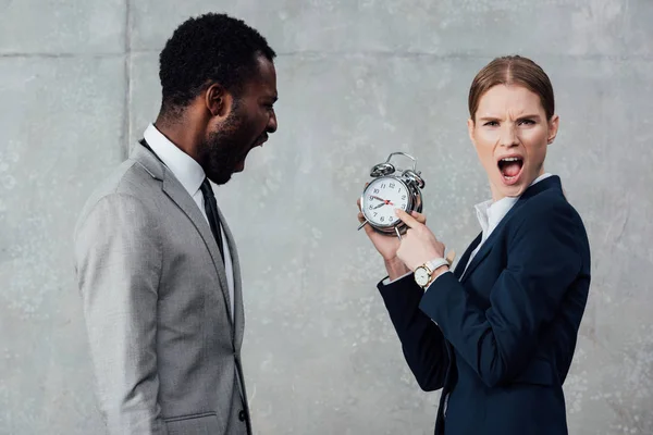 Femme d'affaires regardant la caméra et pointant vers l'horloge tandis que l'homme d'affaires afro-américain criant sur fond gris — Photo de stock