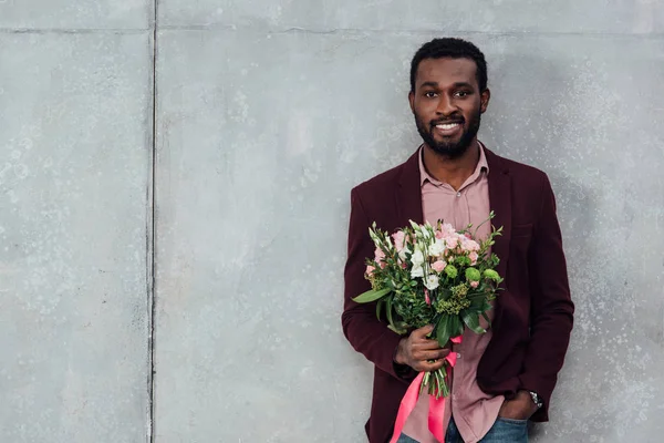 Uomo afroamericano sorridente in abiti casual con fiori e guardando la fotocamera su sfondo grigio — Stock Photo