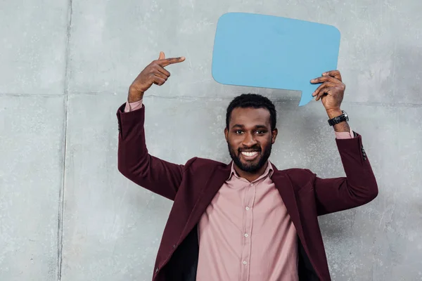 Lächelnder afrikanisch-amerikanischer Geschäftsmann, der in die Kamera schaut und mit dem Finger auf die Sprechblase auf grauem Hintergrund zeigt — Stockfoto