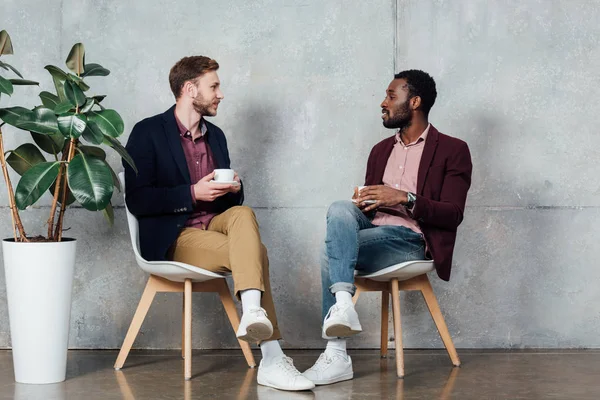 Hombres multiétnicos con ropa casual sentados, tomando café y hablando en la sala de espera — Stock Photo