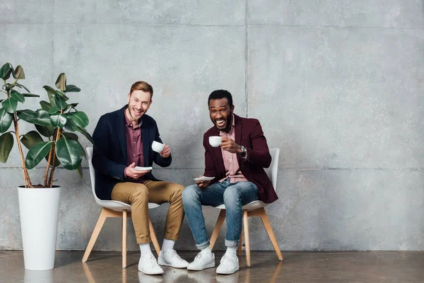 Багатоетнічні чоловіки сидять, дивлячись на камеру і п'ють каву в залі очікування — стокове фото