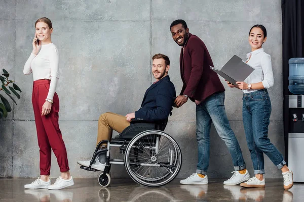 Sorridente gruppo multietnico di persone in abiti casual rotolamento uomo in sedia a rotelle in sala d'attesa — Foto stock