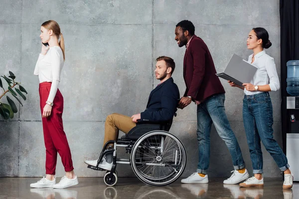 Multiethnische Gruppe von Menschen in Freizeitkleidung rollt Mann im Rollstuhl in Wartehalle — Stockfoto