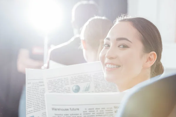 Enfoque selectivo de la mujer de negocios asiática mirando a la cámara y la celebración de periódico con colegas de fondo - foto de stock