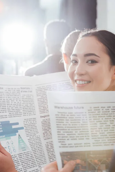 Hermosa asiático mujer de negocios mirando cámara y celebración de periódico con colegas en fondo - foto de stock