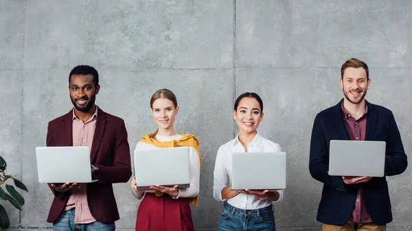 Empresarios casuales multiétnicos que utilizan computadoras portátiles en la sala de espera y mirando a la cámara - foto de stock