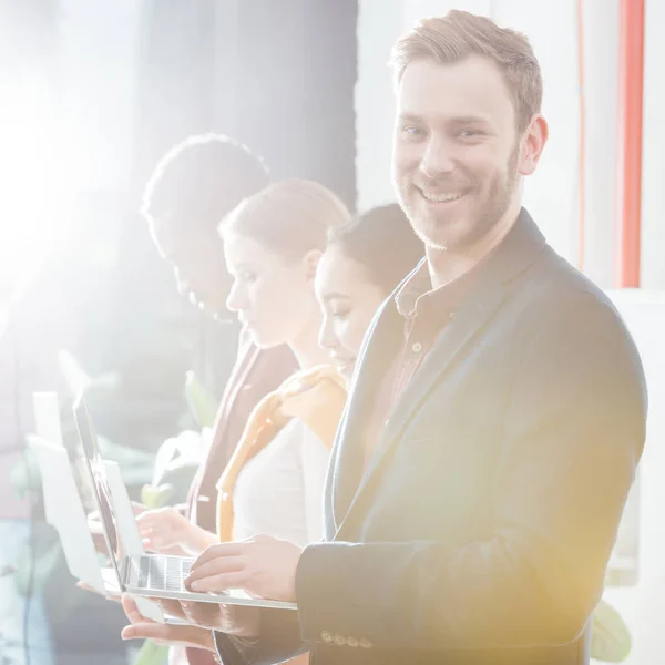 Handsome smiling businessman looking at camera with colleagues using laptops on background — Stock Photo