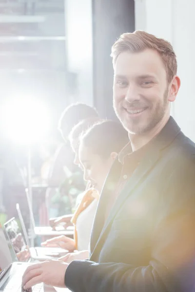 Избирательный фокус красивого улыбающегося бизнесмена, смотрящего в камеру с коллегами, использующими ноутбуки на заднем плане — стоковое фото