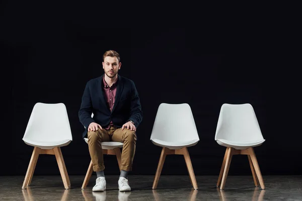 Serious man sitting on chair and looking at camera isolated on black — Stock Photo