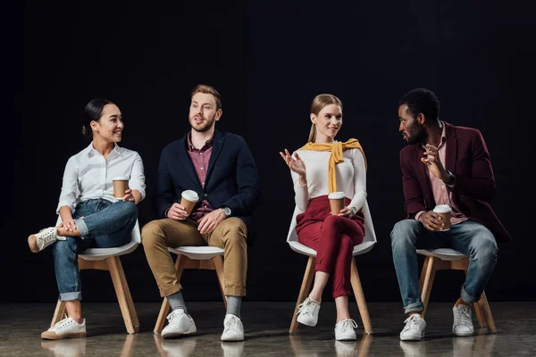 Groupe multiethnique de personnes en vêtements décontractés assis sur des chaises avec du café pour aller isolé sur noir — Photo de stock