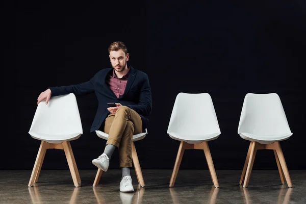 Man sitting on chair, looking at camera and using smartphone isolated on black — Stock Photo