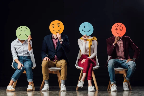 Grupo de pessoas sentadas em cadeiras e segurando cartões faciais com várias emoções isoladas no preto — Fotografia de Stock