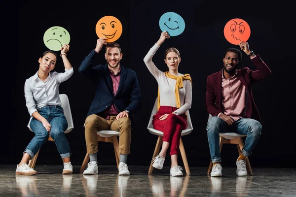 Multiethnic people sitting on chairs and holding face cards with various emotions isolated on black — Stock Photo