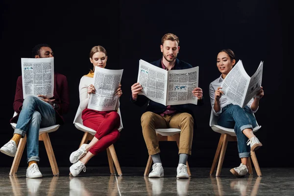 Multiethnische Gruppe fokussierter Gelegenheitsunternehmer, die auf Stühlen sitzen und isoliert auf schwarz Wirtschaftszeitungen lesen — Stockfoto