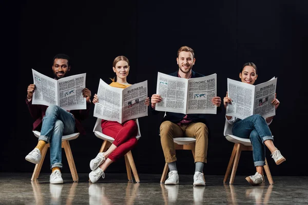 Sonriente grupo multiétnico de empresarios casuales sentados en sillas y leyendo periódicos de negocios aislados en negro - foto de stock