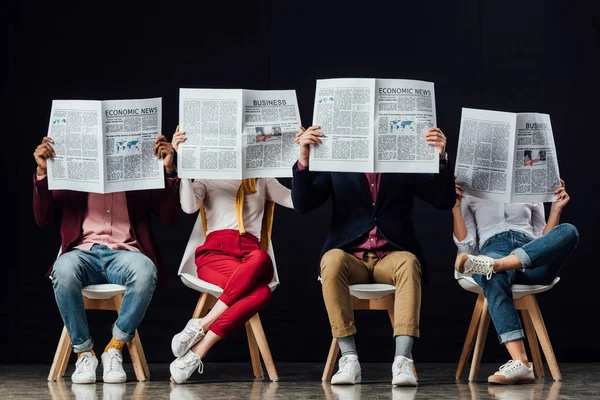 Gruppe von Gelegenheitsunternehmern mit obskuren Gesichtern, die auf Stühlen sitzen und isoliert auf schwarz Wirtschaftszeitungen lesen — Stockfoto