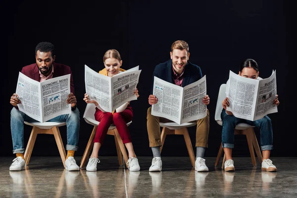 Multiethnic group of casual businesspeople sitting on chairs and reading business newspapers isolated on black — Stock Photo