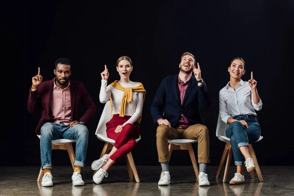 Multiethnic group of people sitting on chairs and showing idea gestures isolated on black — Stock Photo