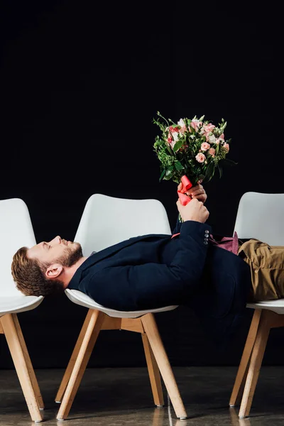 Uomo con gli occhi chiusi sdraiato su sedie e tenendo fiori isolati su nero — Foto stock