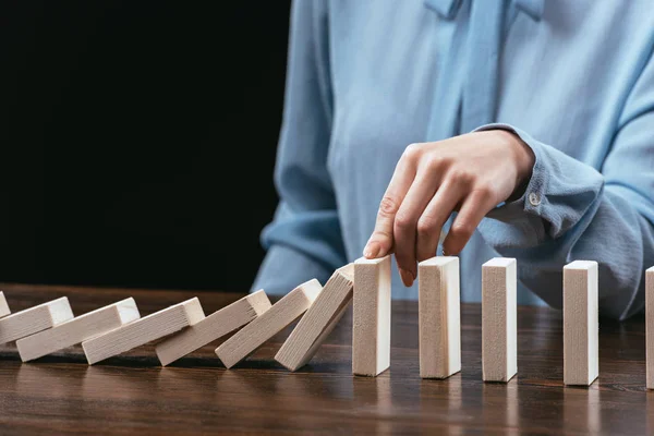 Partial view of woman sitting at desk and preventing wooden blocks from falling isolated on black — Stock Photo