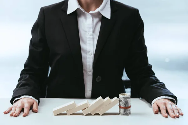 Cropped view of businesswoman sitting at table with fallen row of wooden blocks and money roll — Stock Photo
